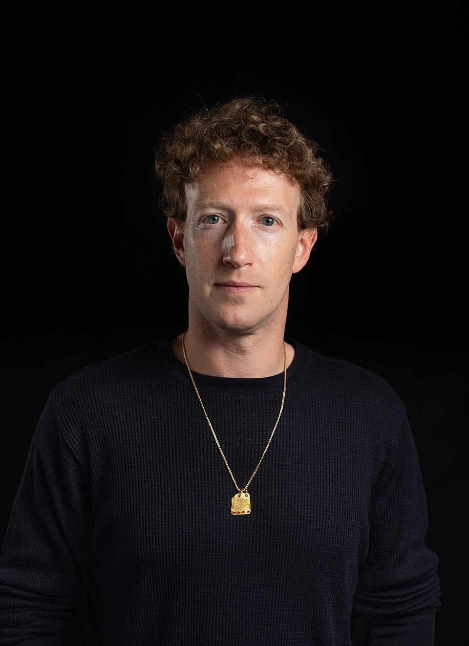 Mark Zuckerberg wearing a dark sweater and gold necklace against a black background