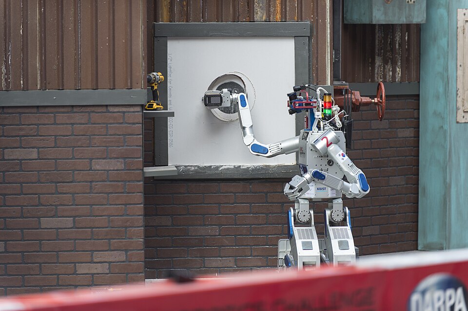 A white and blue humanoid robot holding a power tool against a white wall panel on a brick-walled competition course.