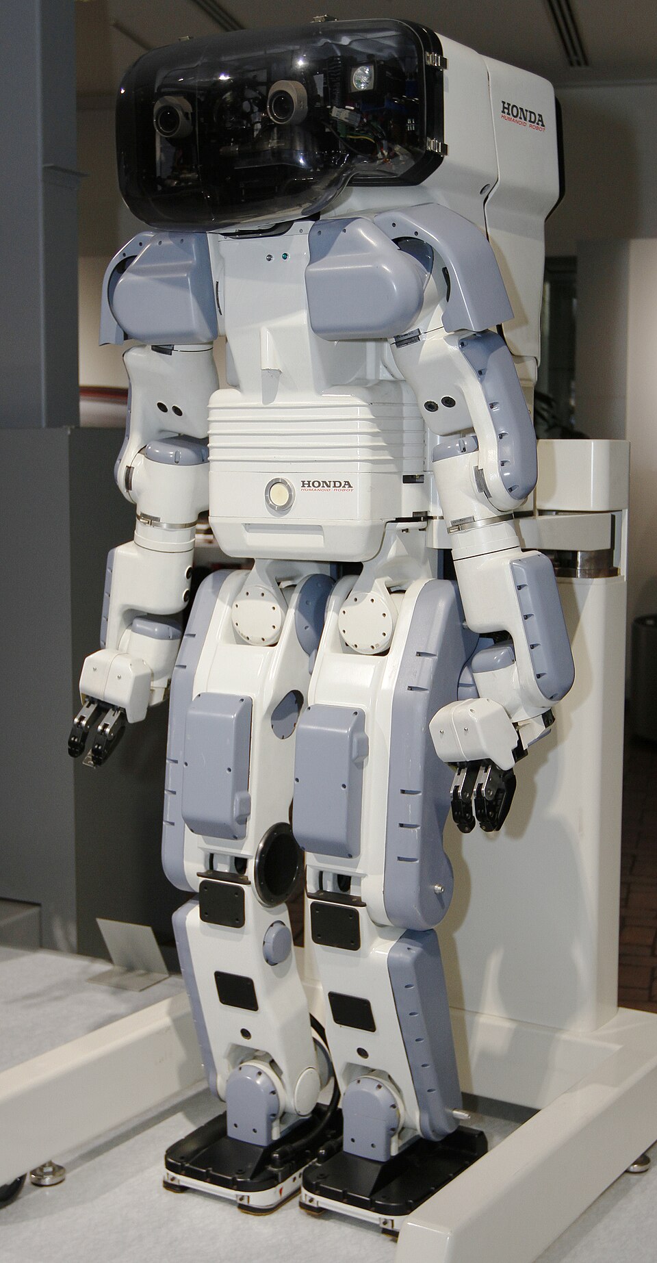 A tall white and gray humanoid robot with a large box-shaped black head unit and a backpack, standing on a museum platform.