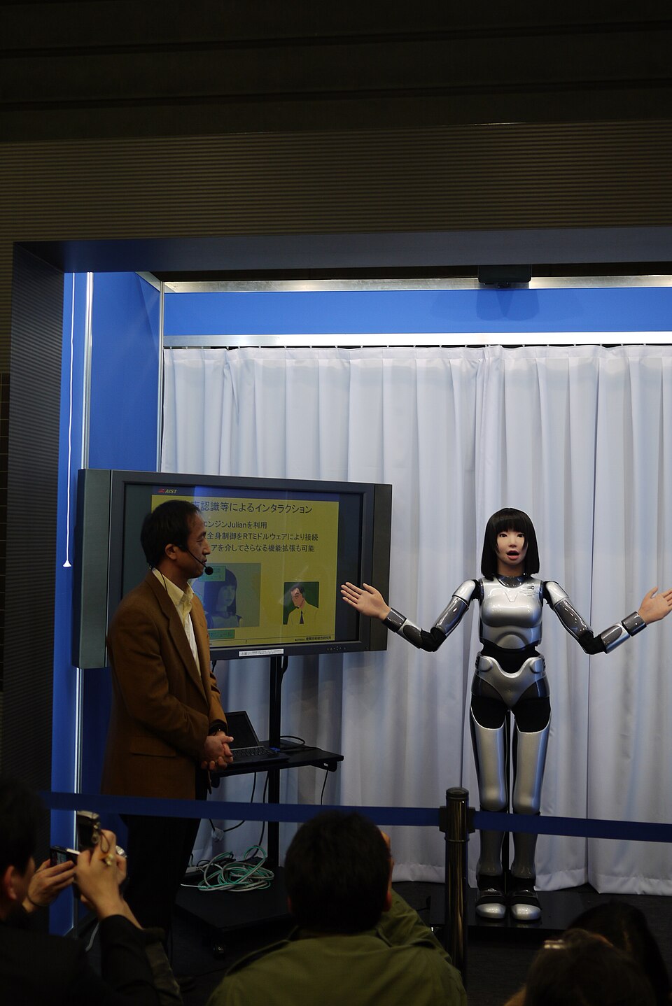 A slim silver humanoid robot with a lifelike female face and black hair, arms spread, on a small stage beside a presenter.
