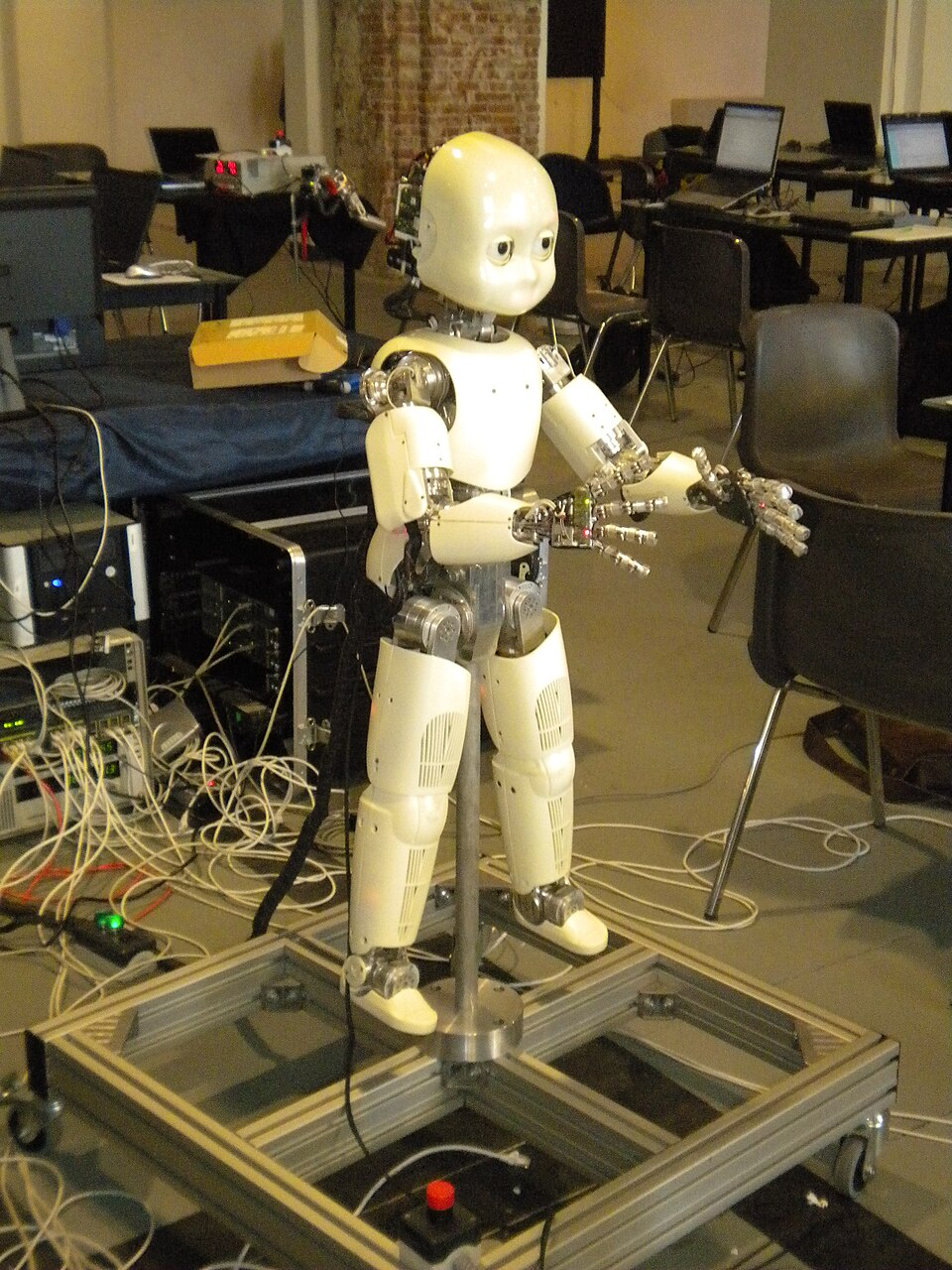 A child-sized white and silver humanoid robot with a cartoonish face standing on a metal support frame in a laboratory.