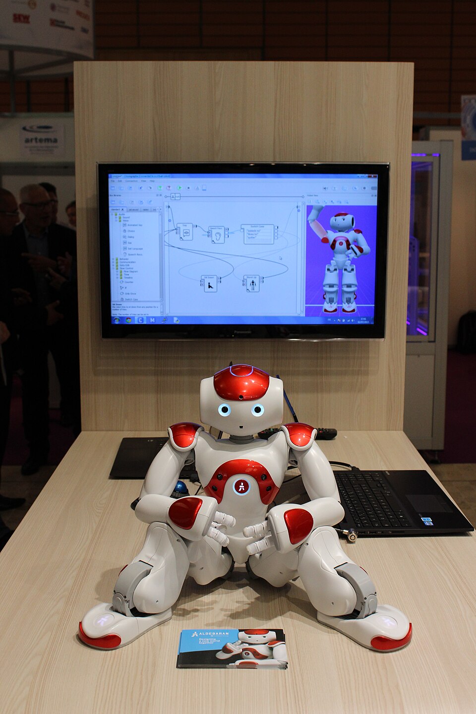 A small white and red humanoid robot sitting on a table in front of a monitor showing programming software.
