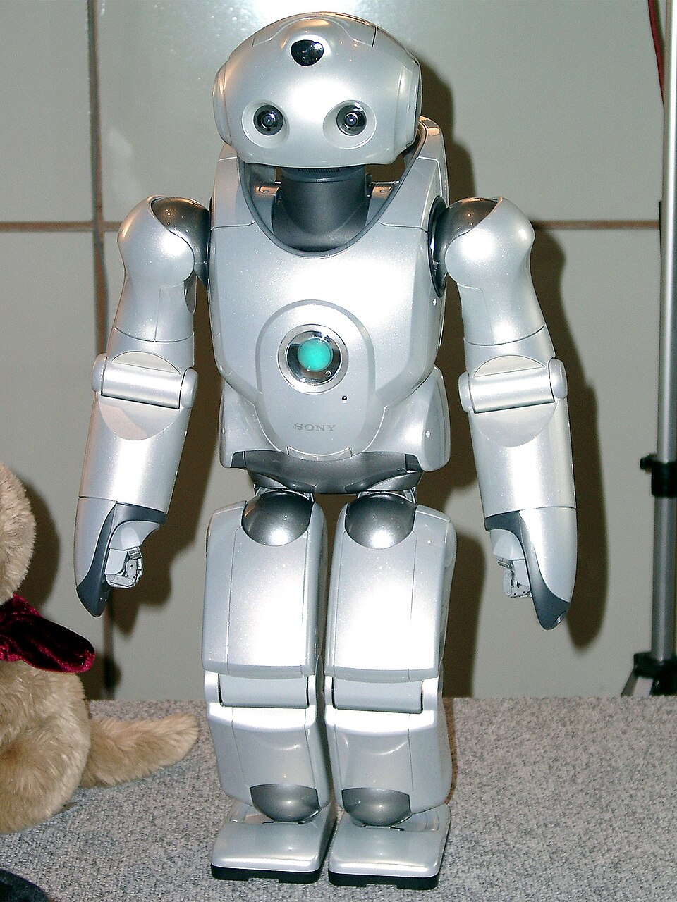 A small silver humanoid robot with a round head and a glowing chest light standing on a gray carpeted table.