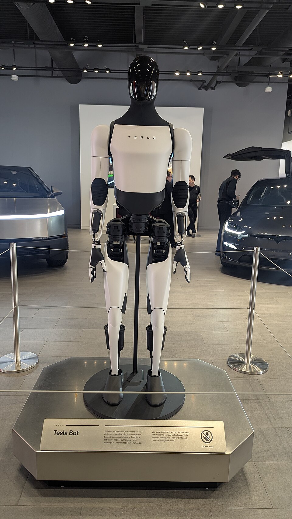 A white and black humanoid robot standing on a display plinth inside a car showroom, with vehicles in the background.