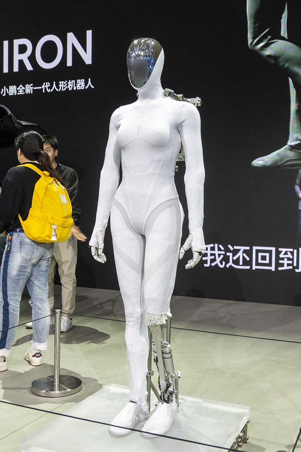 A slender white humanoid robot with a humanlike body shape and glossy faceless head, one lower leg uncovered to show metal parts.