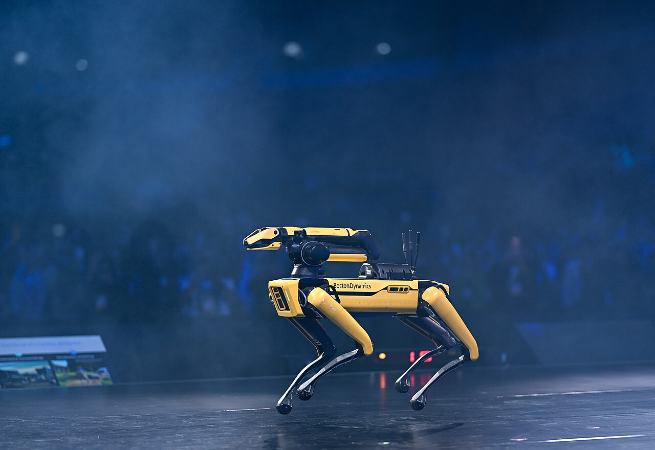 Yellow-and-black Boston Dynamics Spot robot with a robotic arm on its back on stage.