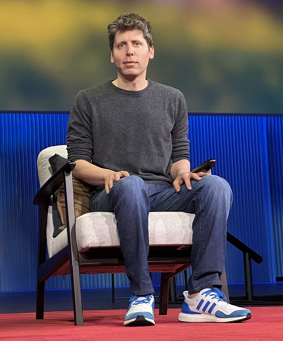 Sam Altman seated on stage wearing a grey shirt and headset microphone.