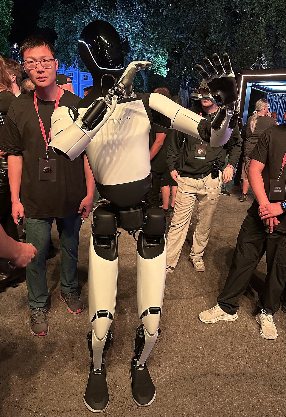 A white-and-black Tesla Optimus robot with raised hands at an October 2024 event.