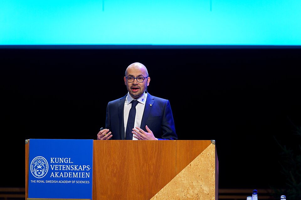 Demis Hassabis speaking behind a lectern in a dark suit and glasses
