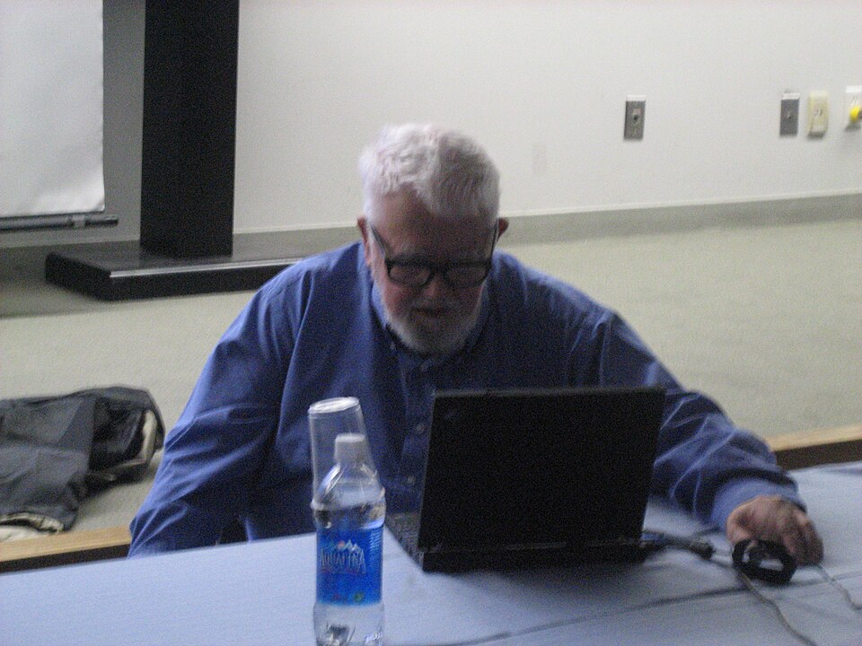 John McCarthy seated behind a laptop, wearing glasses and a blue shirt
