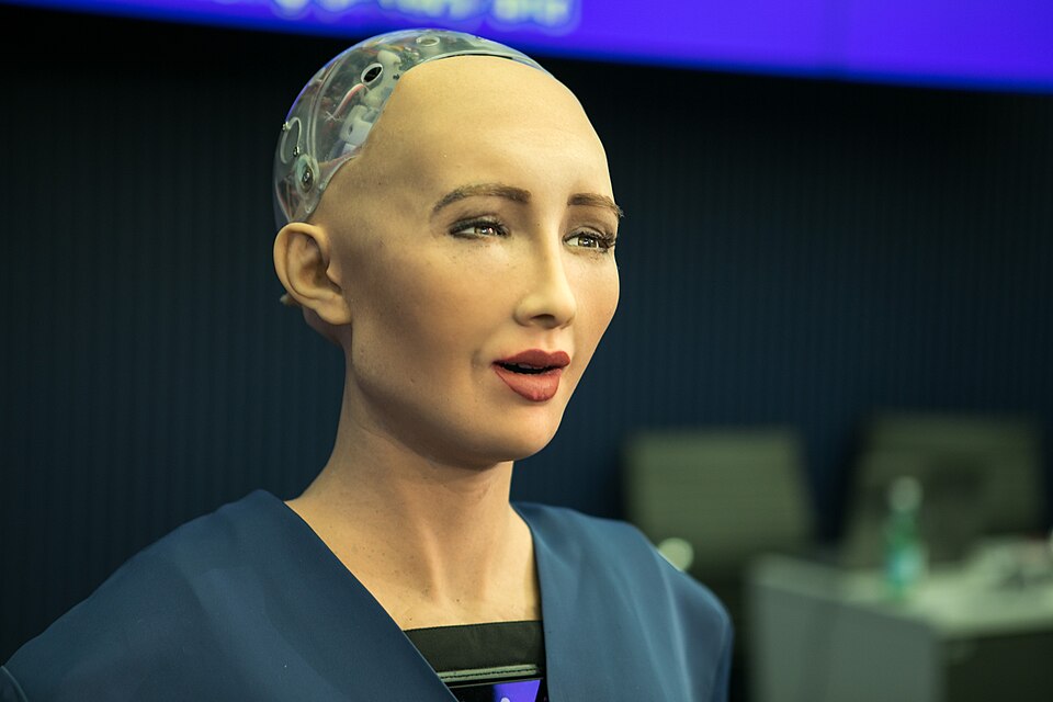 Sophia robot in blue clothing, with a humanlike face and exposed clear upper head