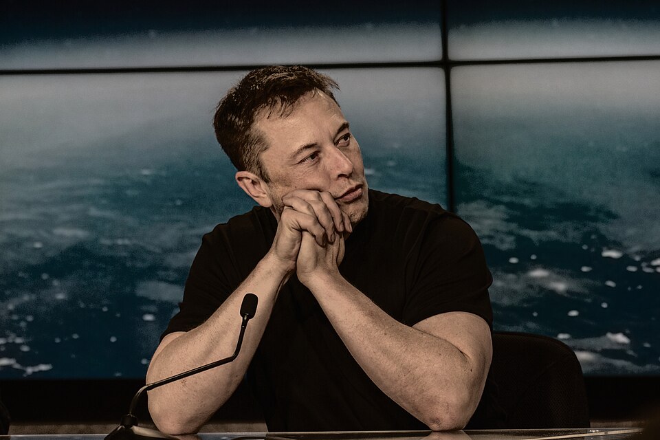 Elon Musk seated at a microphone, wearing a black shirt with his hands clasped beneath his chin