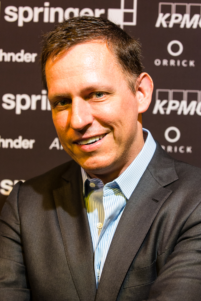 Peter Thiel wearing a dark suit jacket and a blue striped shirt in front of an event backdrop